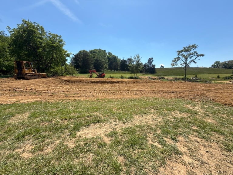 Rural farmland with cleared brown soil, green pasture, scattered trees, and construction equipment under a clear blue sky