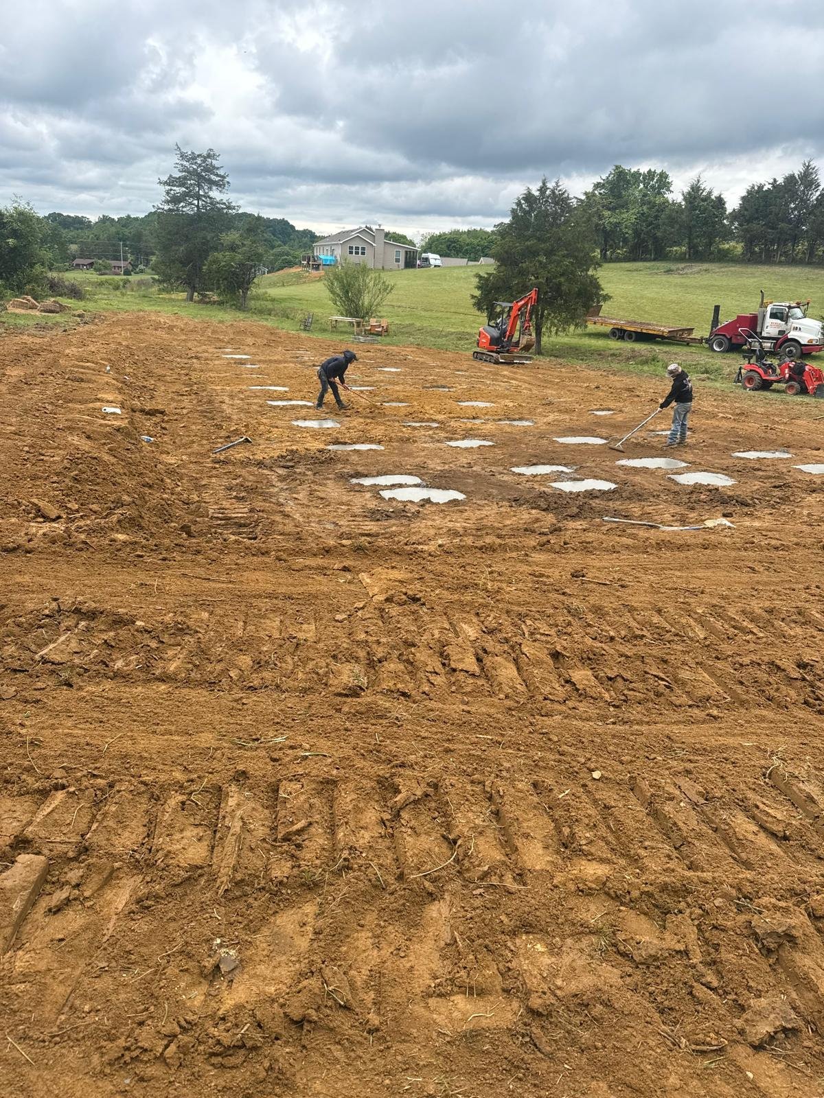 Two workers preparing a large dirt field with heavy machinery visible in the background, including a red excavator and tractor, on an overcast day