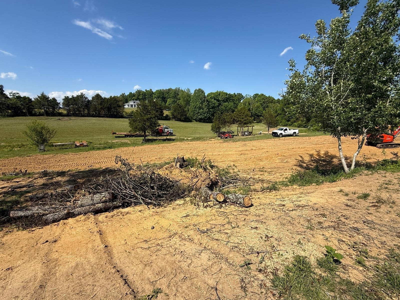 Cleared rural land with logs, heavy machinery, and trees under blue sky