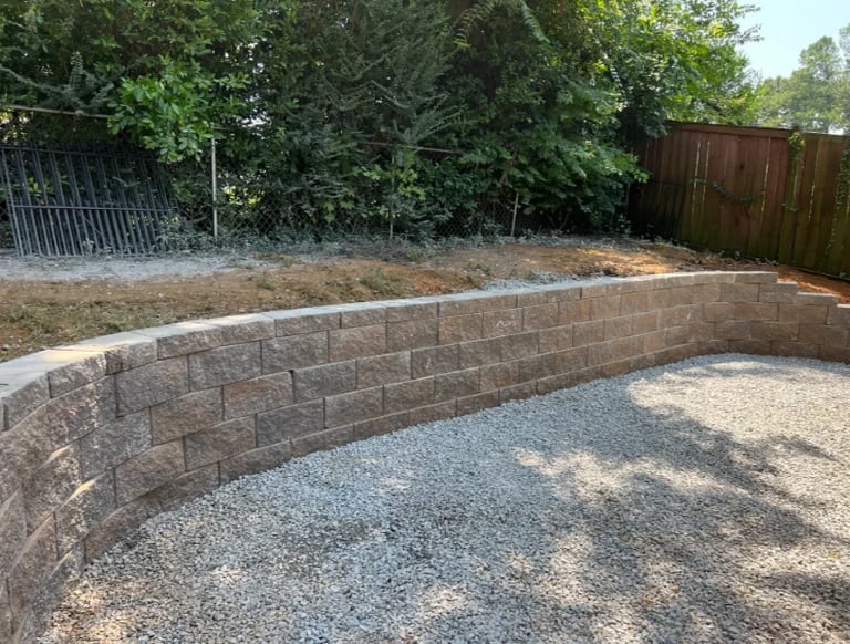 Curved stone retaining wall with gravel base in residential backyard surrounded by trees and wooden fence