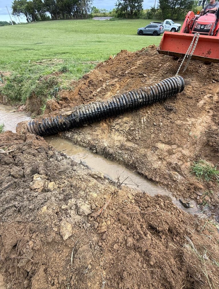 Black corrugated drainage pipe laid in muddy earth with red truck and green field in background