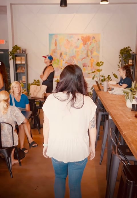 Woman in white shirt and jeans stands in an indoor event space with guests and plants