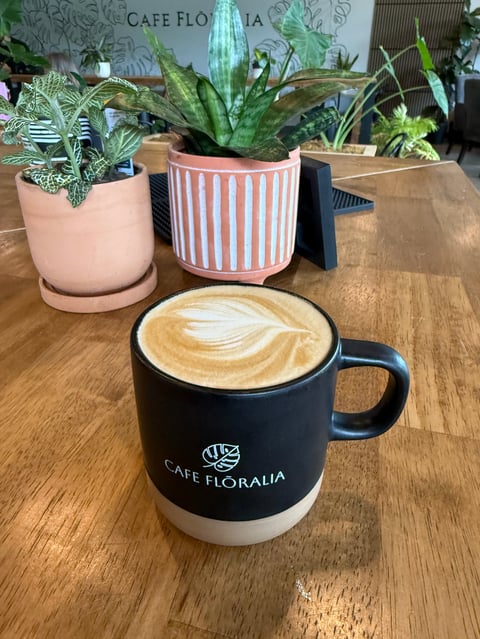 Black Cafe Floralia mug with latte art on wooden table surrounded by potted plants