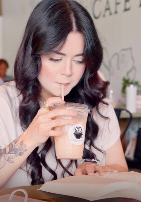 Woman with long dark wavy hair drinking from a coffee cup at a cafe table while reading