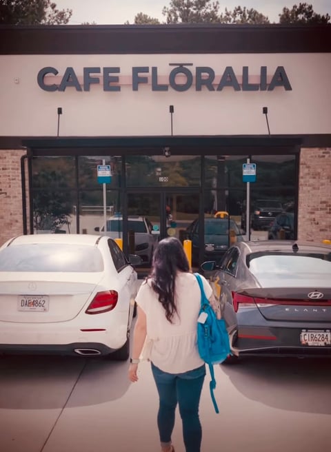 Woman with blue backpack walking toward Cafe Floralia storefront in a parking lot