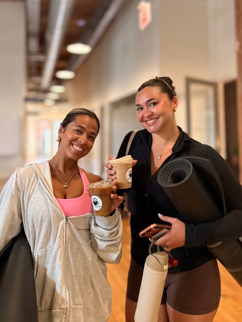 Two smiling women holding coffee cups and yoga mat in an indoor hallway