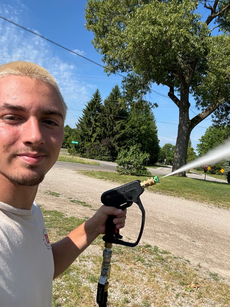 Man holding a garden hose sprayer on a rural road with trees and blue sky in the background