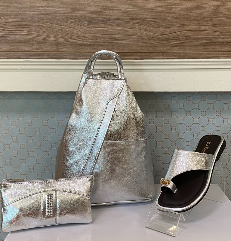 Silver metallic backpack, clutch, and sandal displayed on a table with wood paneling and textured blue wallpaper background