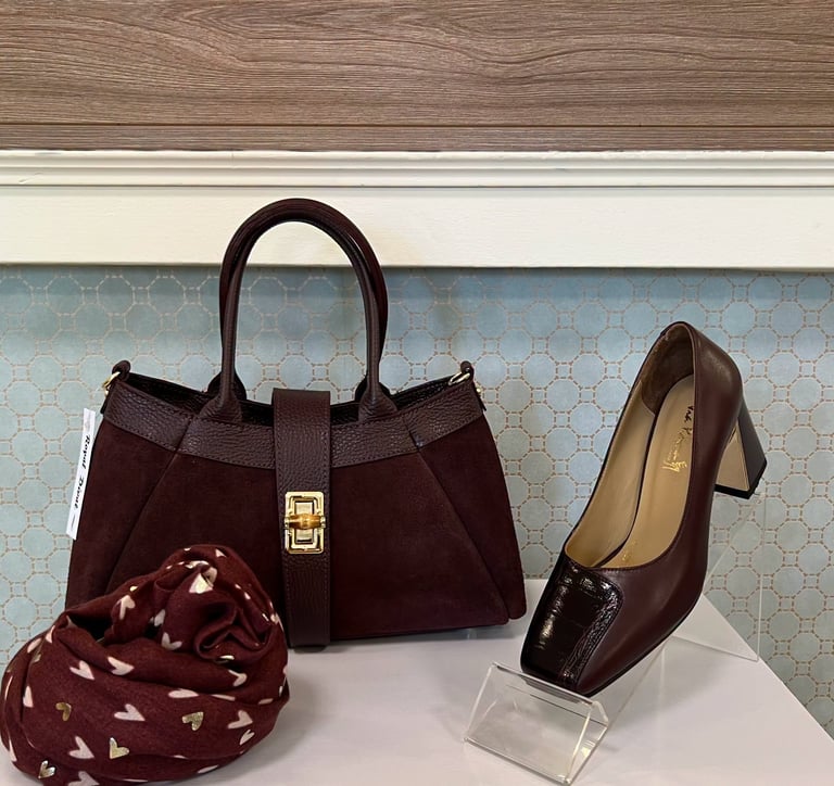 Burgundy leather tote bag, brown block heel shoe, and patterned scarf displayed on white surface against patterned wallpaper