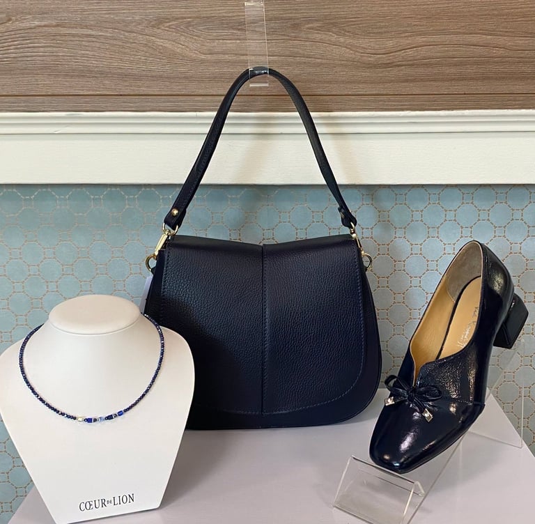 Navy blue shoulder bag displayed with white straw hat and black leather loafers on a light blue geometric patterned backdrop