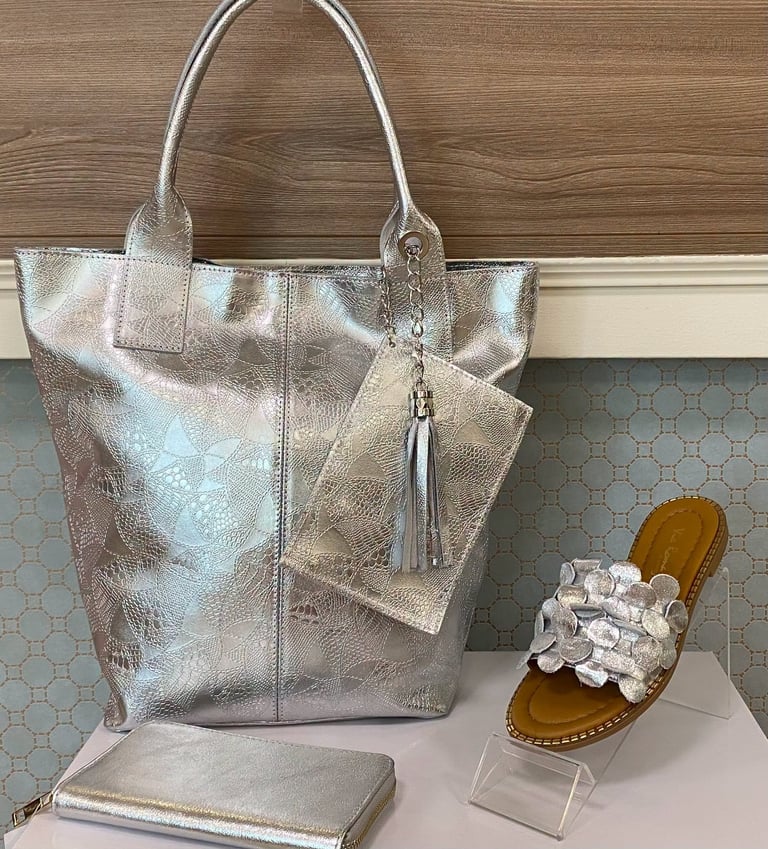 Silver metallic tote bag with matching wallet and key charm displayed with white ruffled sandal on wooden background