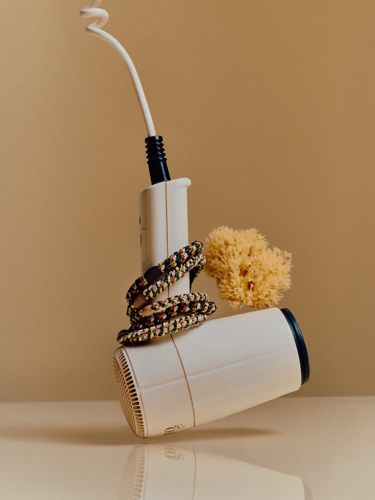 Two white and gold hair styling tools stacked together with a patterned cord and yellow flower against a beige background