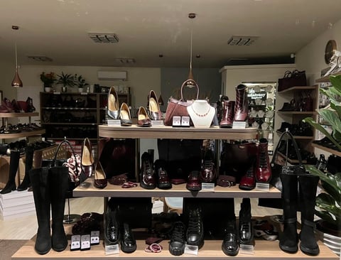 A retail shoe store display with organized shelves showing various black shoes, boots, and accessories with pendant lighting and decorative plants