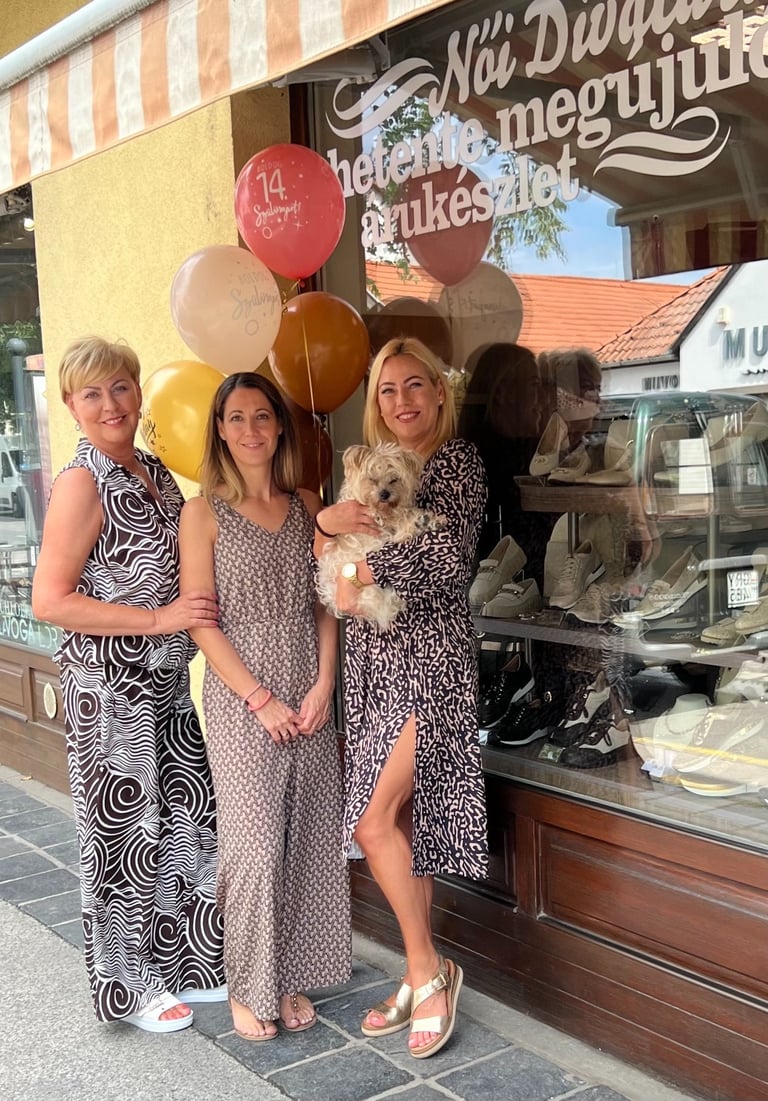 Three women and a small dog posing outside a storefront with red and beige balloons