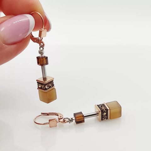 Hand holding geometric crystal drop earrings with rose gold components and amber-colored cube beads
