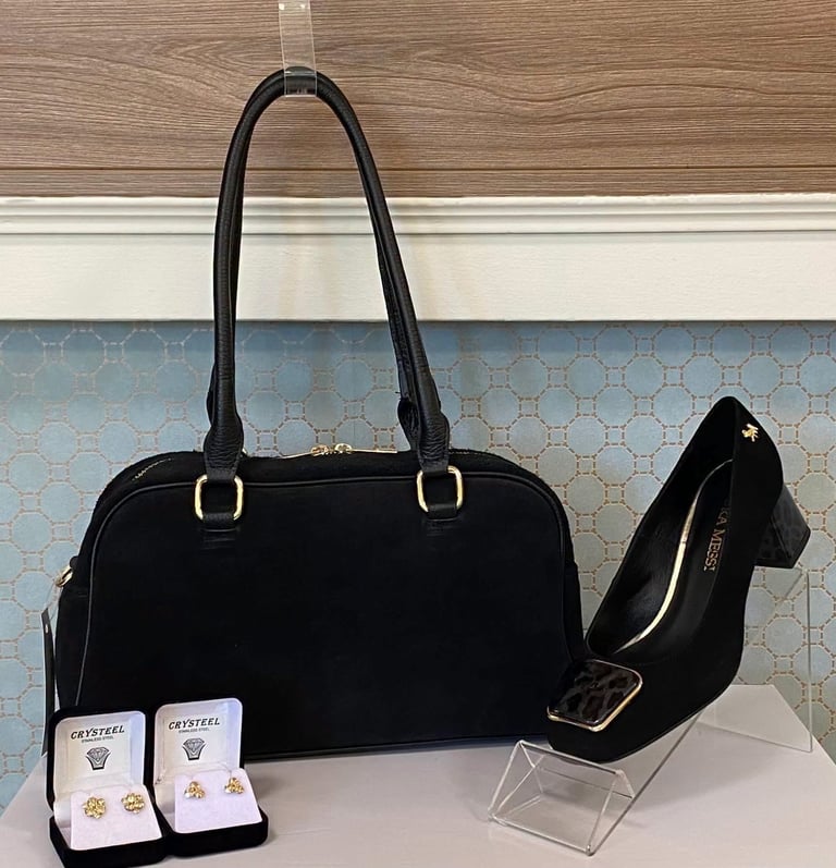 Black handbag with gold hardware displayed next to black heeled shoe and jewelry boxes on white surface