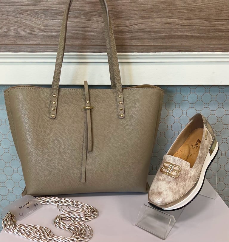 Beige leather tote bag with studded handles displayed next to a suede loafer shoe and striped cord on a light background