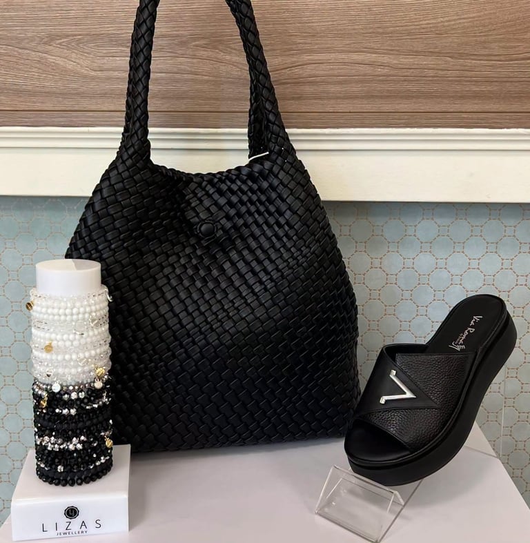 Black woven tote bag displayed with matching black slides and decorative beaded bracelets on white surface