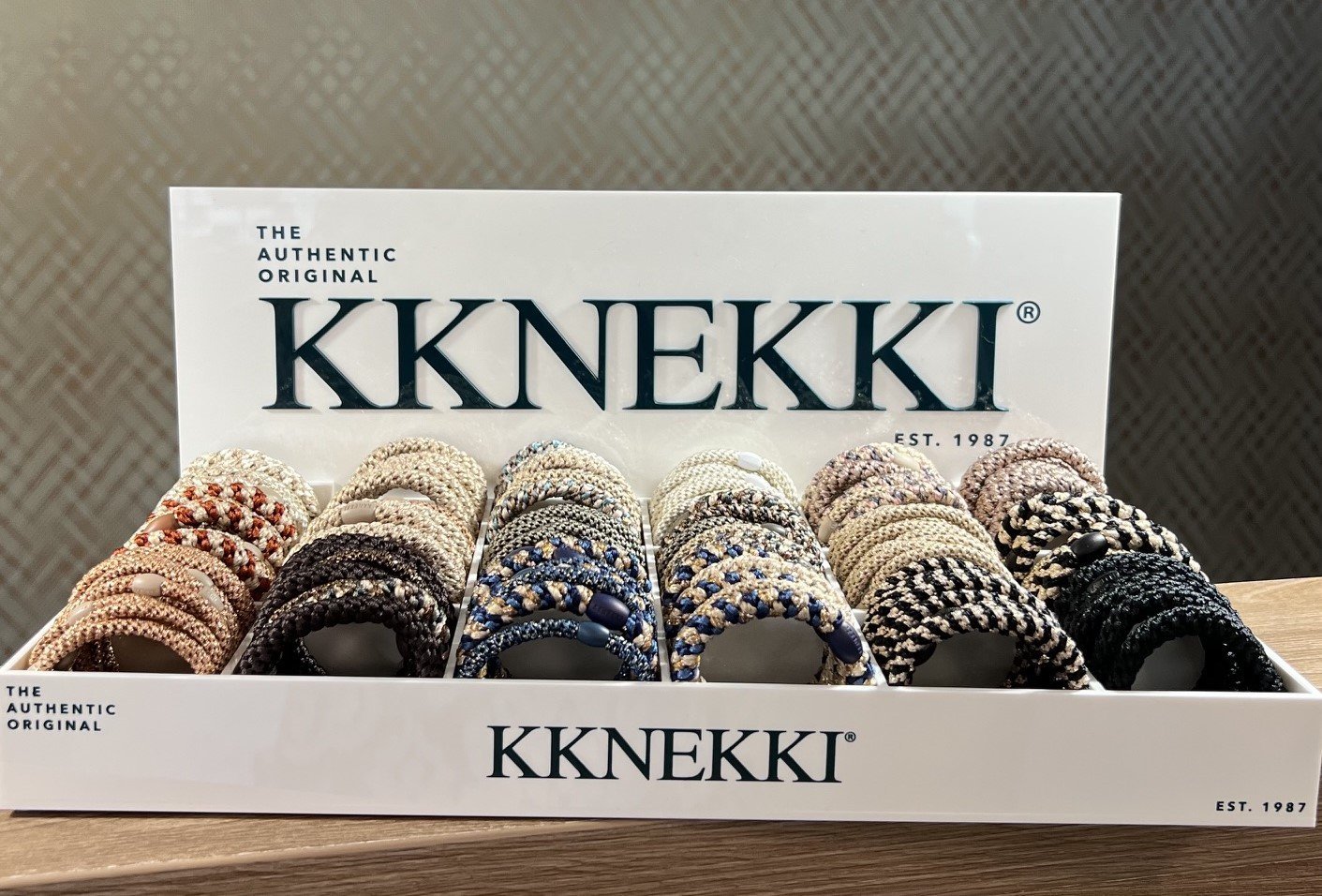 Display case of KKNEKKI hair coils in various colors including gold, black, navy, gray, and beige arranged in rows