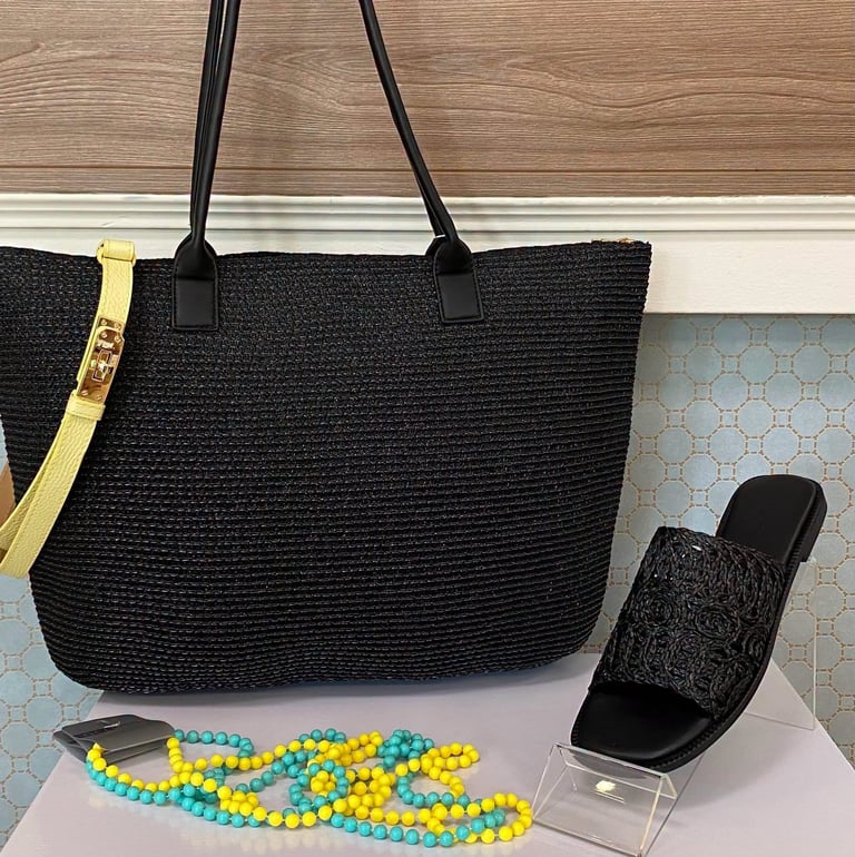 Black woven tote bag with leather handles displayed on white shelf, alongside colorful beaded necklace and black slide sandal