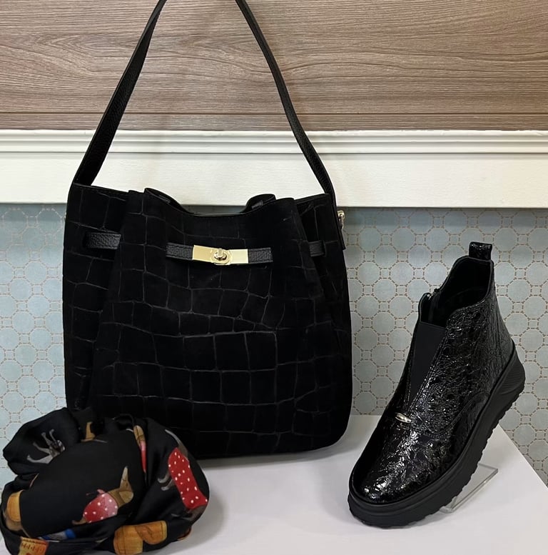 Black crocodile-textured handbag with shoulder straps displayed on white shelf alongside black glittery ankle boot and decorative beaded item