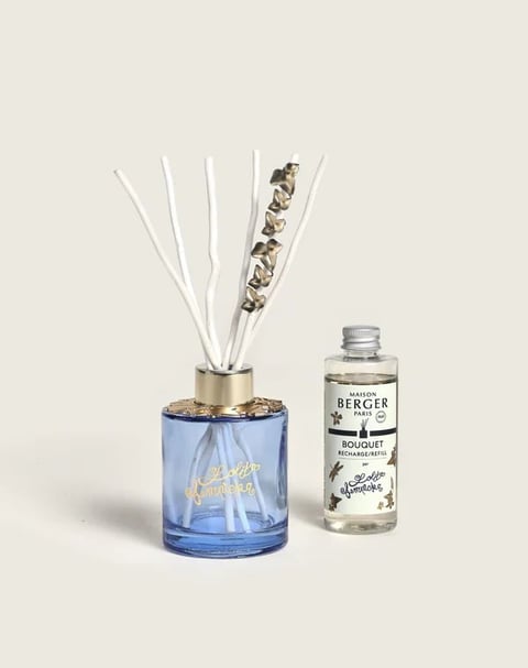 A blue room diffuser with decorative reeds and a clear refill bottle labeled Maison Berger on a neutral background