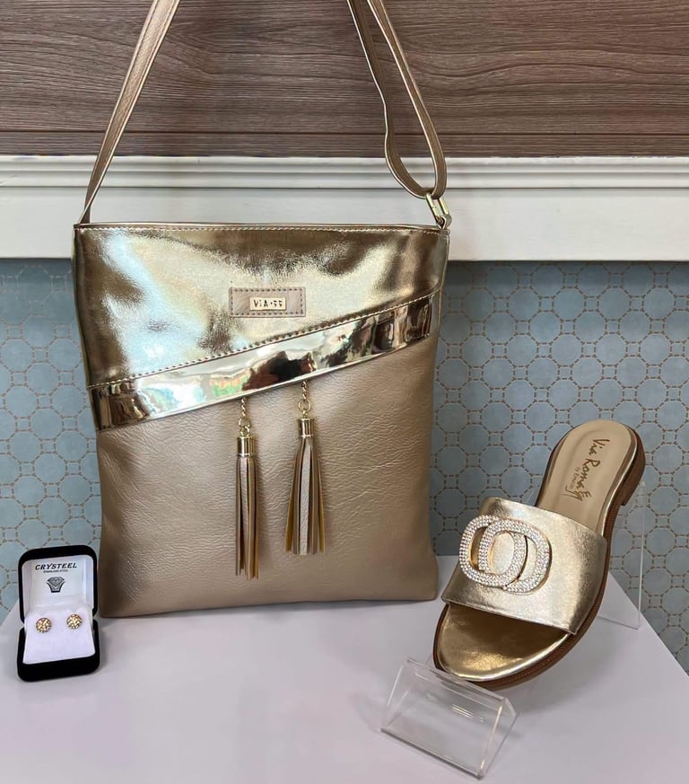 Gold metallic crossbody bag with tassel detail next to matching slide sandal and smartphone displaying product information