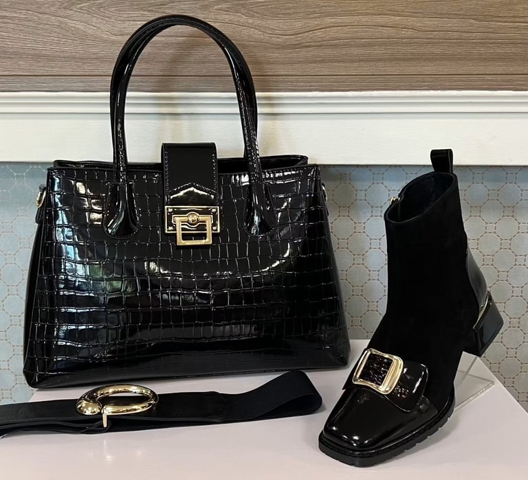 Black crocodile-textured handbag with gold clasp, matching ankle boot, black belt, and gold bangle displayed on white shelf