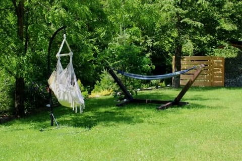 Backyard garden with white macramé hanging chair, hammock with stand, and wooden pergola surrounded by green trees and lawn
