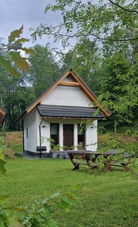 Small white cottage with brown roof and porch, wooden picnic table in green garden surrounded by trees