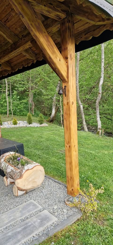Wooden pergola post in a backyard with green lawn, forest backdrop, and concrete stepping stones in foreground