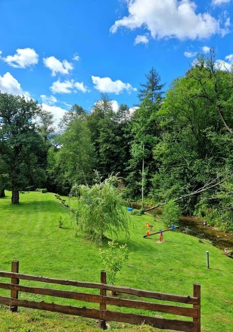 Scenic pastoral landscape with green meadow, wooden fence, stream, and dense trees under blue sky with white clouds