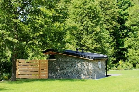 Modern stone cabin with solar panels on roof, wooden fence, surrounded by lush green trees and grass