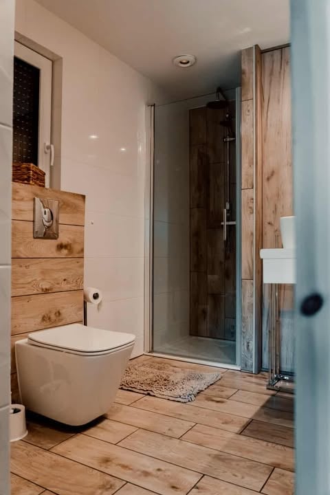 Modern bathroom with wooden flooring, white toilet with wooden accent wall, and glass shower enclosure with dark tiles