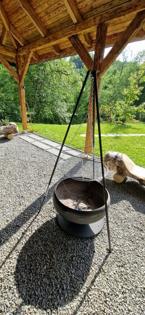 Black cauldron suspended by metal tripod under wooden pergola, gravel patio with green lawn and trees in background