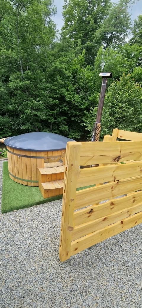 Wooden hot tub with metal cover and chimney surrounded by lush green trees and wooden fence gate