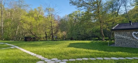 Scenic backyard with lush green lawn, stone pathway, trees, and a small stone building on the right