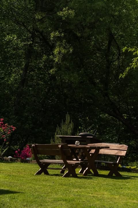 Wooden picnic table on green lawn with pink flowers and dense green trees in background