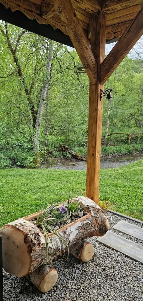 Log planter with purple flowers under wooden pergola overlooking wooded yard with green grass and stream
