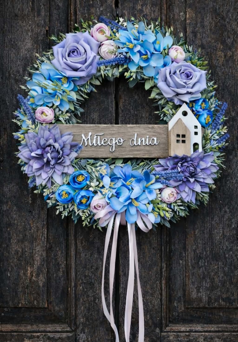 Decorative floral wreath with blue and purple flowers, white house ornament, and Miłego dnia text on wooden door