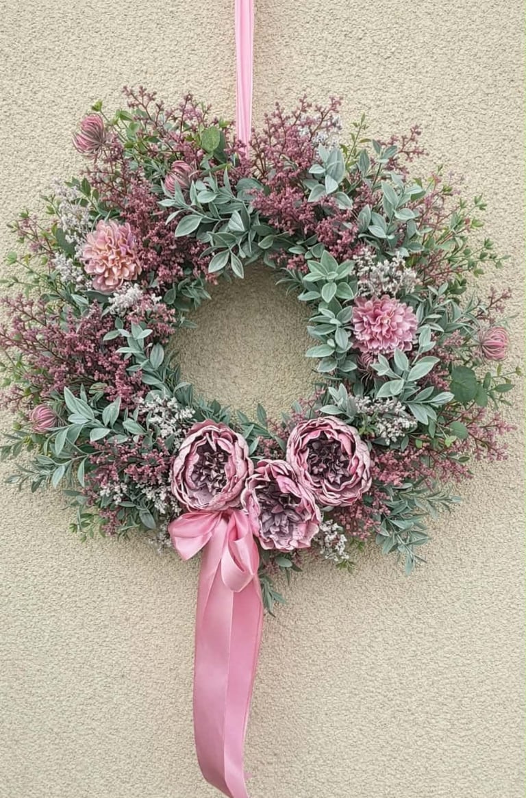 Decorative floral wreath with pink peonies, dusty miller foliage, and small pink flowers tied with a pink ribbon bow, hanging on a beige wall