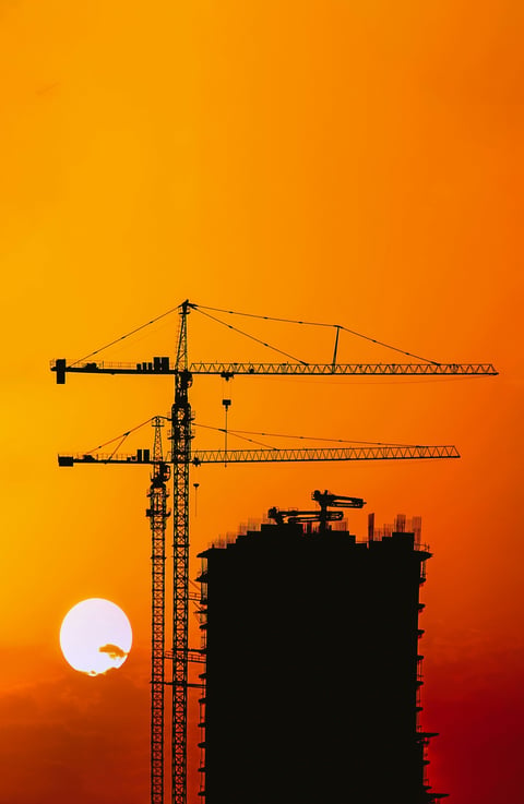Silhouette of construction cranes and building against orange sunset with moon on orange sky