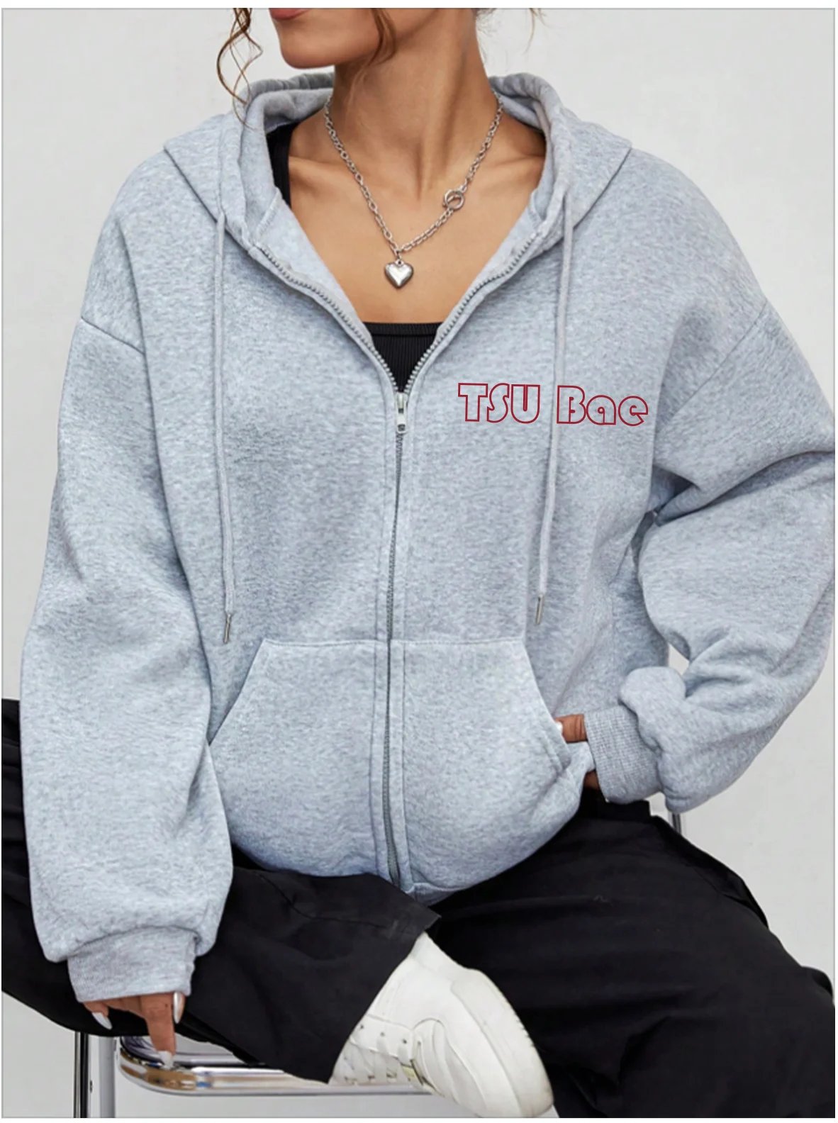 Woman wearing a light gray TSU Bae zip-up hoodie with black pants and a heart pendant necklace