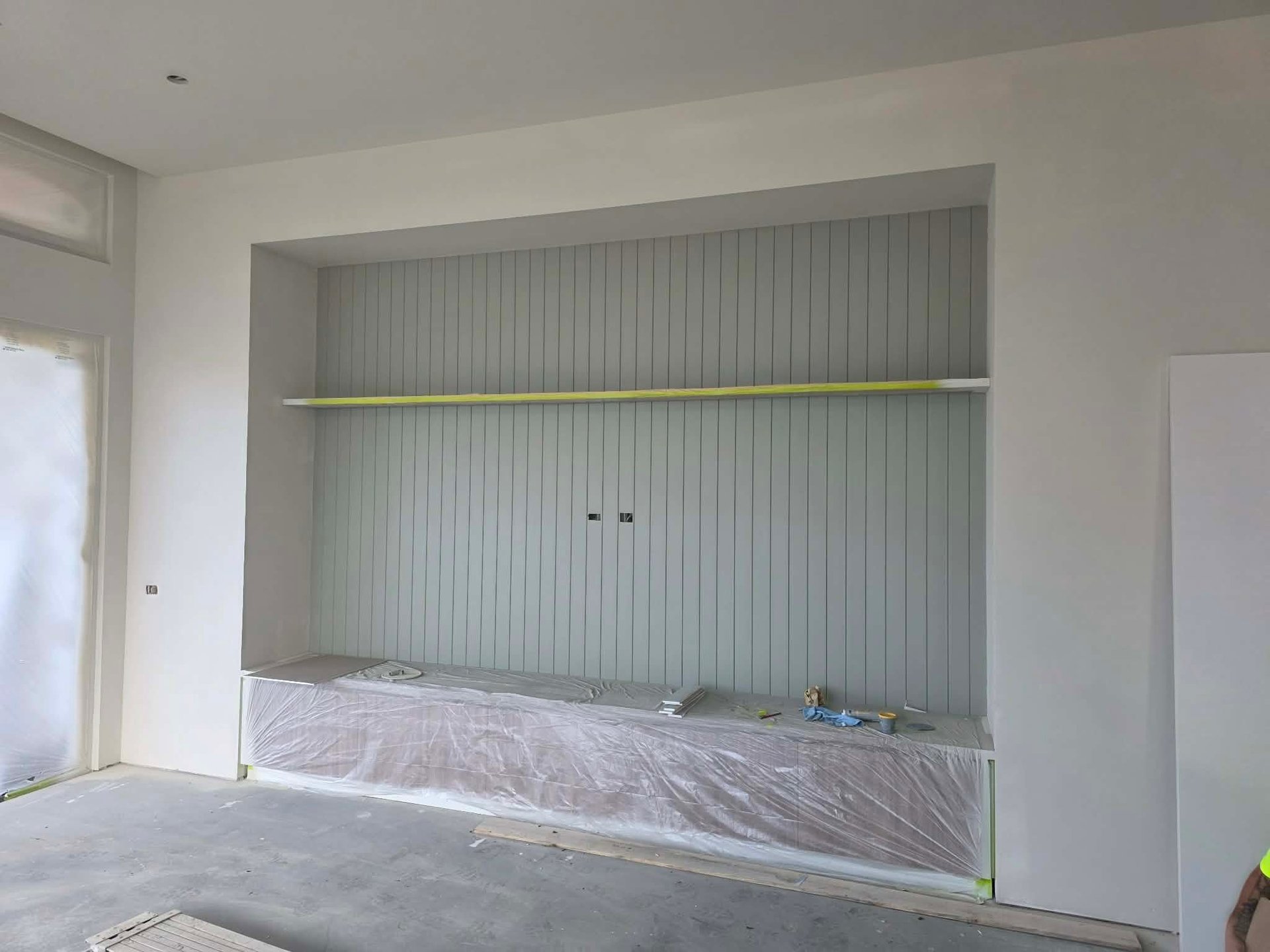 Empty room under construction with white walls, plastic sheeting on floor, and recessed wall niche with metal shelf and vertical louvers