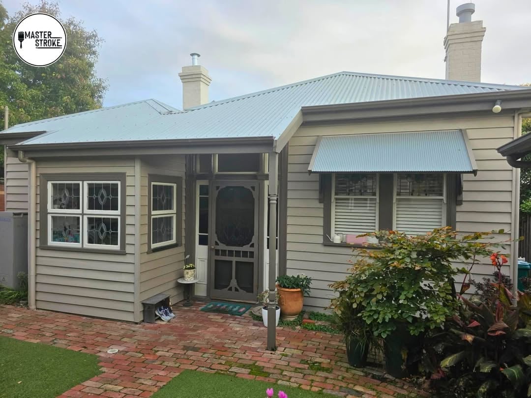 Cream-colored cottage with blue metal roof, bay window, and brick walkway surrounded by green gardens and landscaping