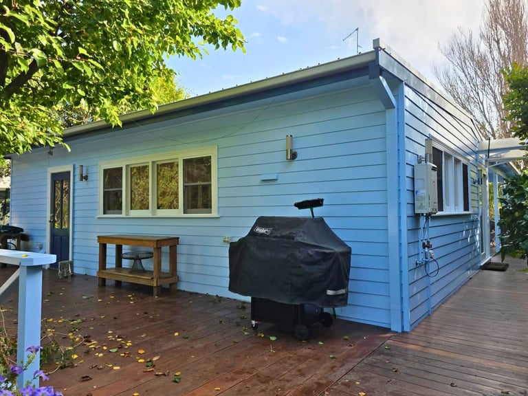 Light blue modern house with large windows, covered porch, and black grill on deck surrounded by green foliage