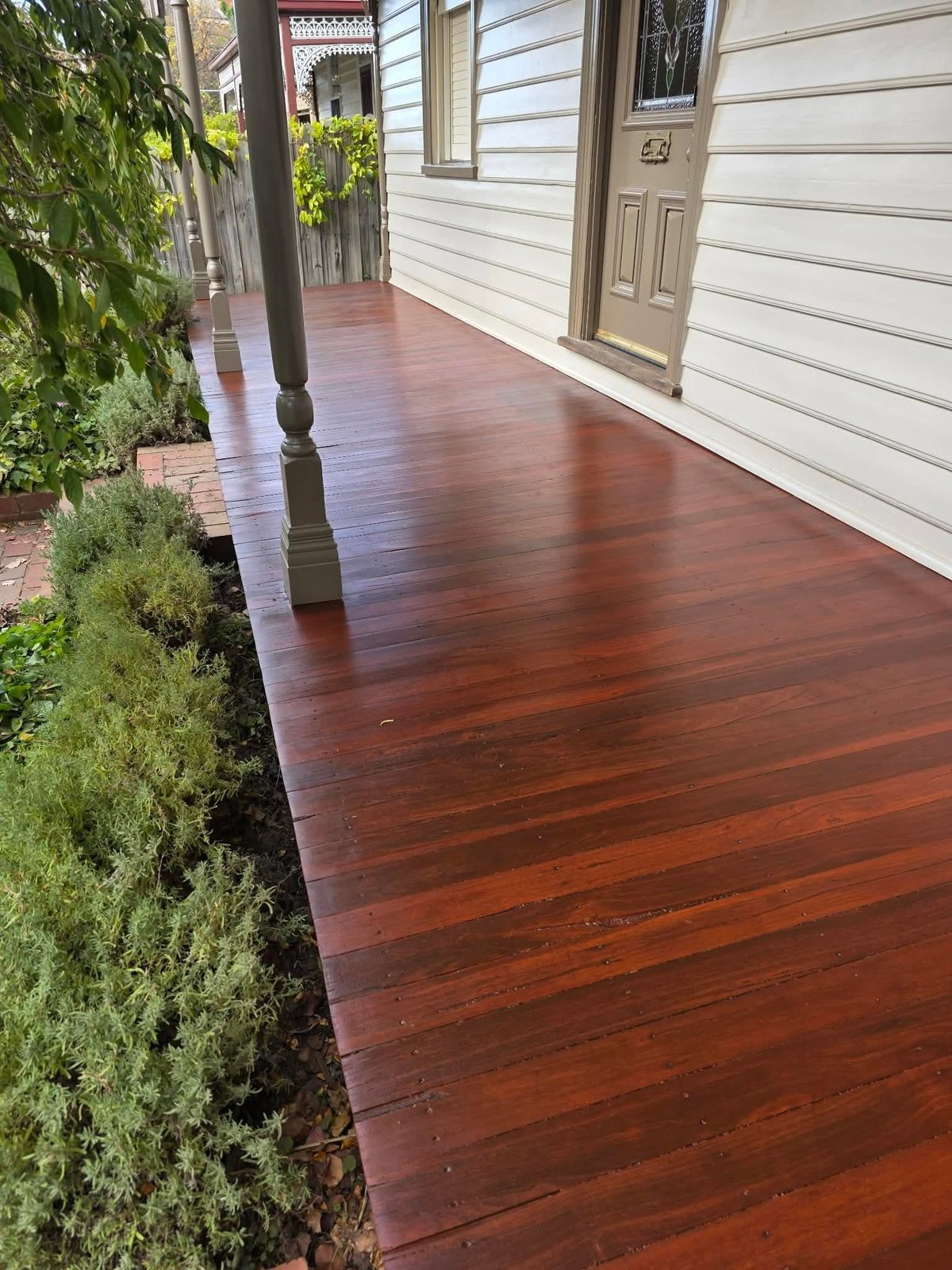 Wooden porch with rich reddish-brown stain, white house siding, beige door, and green plants along the edge on a sunny day