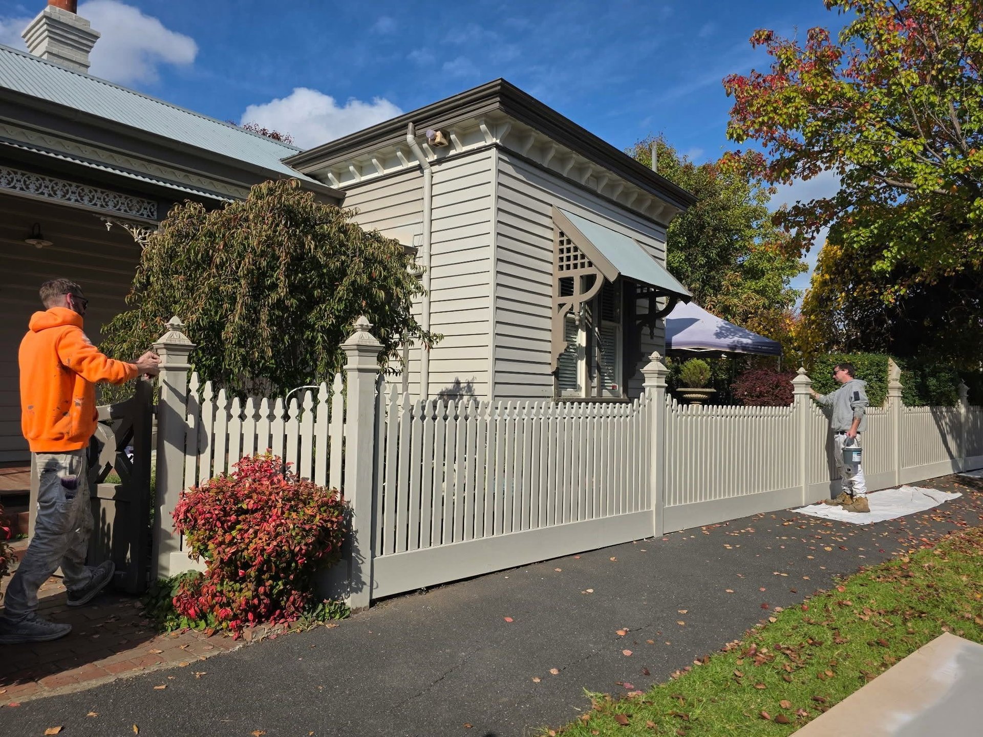 Victorian-style house with white picket fence, red flowering shrubs, and two people on the driveway on a sunny day