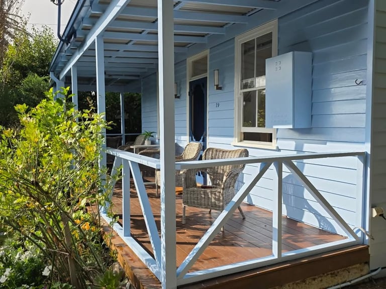 Light blue cottage with covered porch, wooden deck, and white railings, surrounded by green shrubs