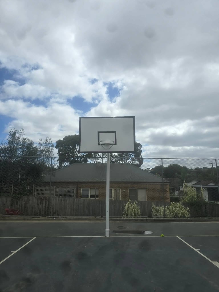 A basketball hoop and backboard stand on an outdoor court with a brick house and cloudy sky in the background